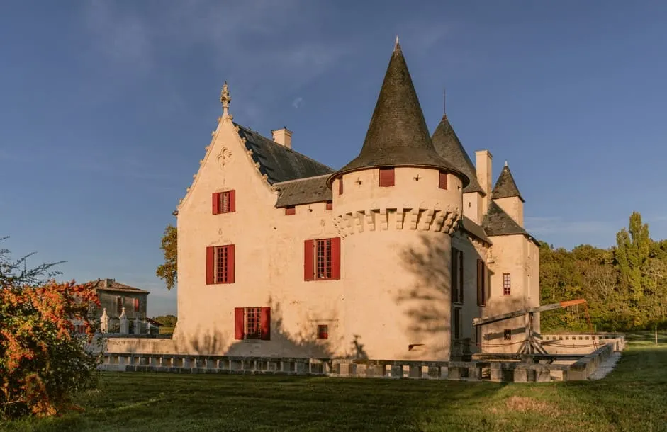 Château Olivier à Bordeaux, château de style médiéval aux murs roses avec tours coniques coiffées de toits pointus et fenêtres rouges, entouré de verdure et d'un parc.