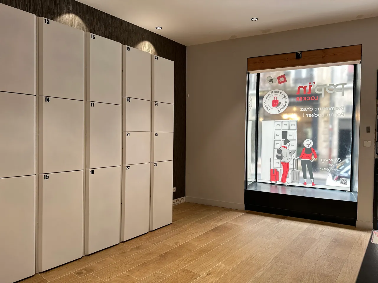 Intérieur d'un espace Popin Locker à Bordeaux avec des casiers de stockage numérotés en crème sur le mur gauche et une vitrine publicitaire illuminée sur le mur droit, sol en parquet clair.