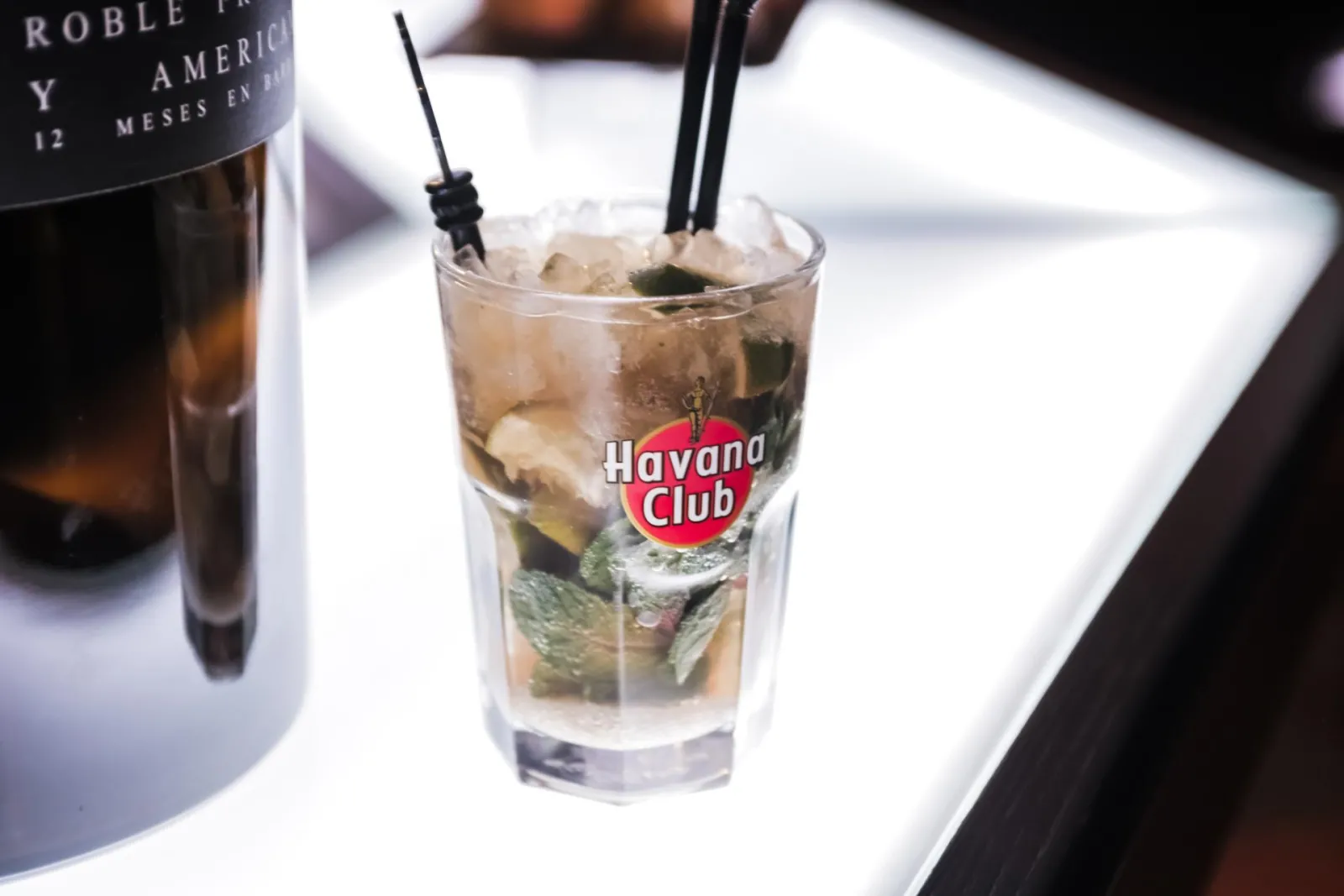 Un verre de cocktail Havana Club avec glaçons et pailles noires, accompagné d'une bouteille de rhum vieilli, présenté dans un cadre de bar élégant.