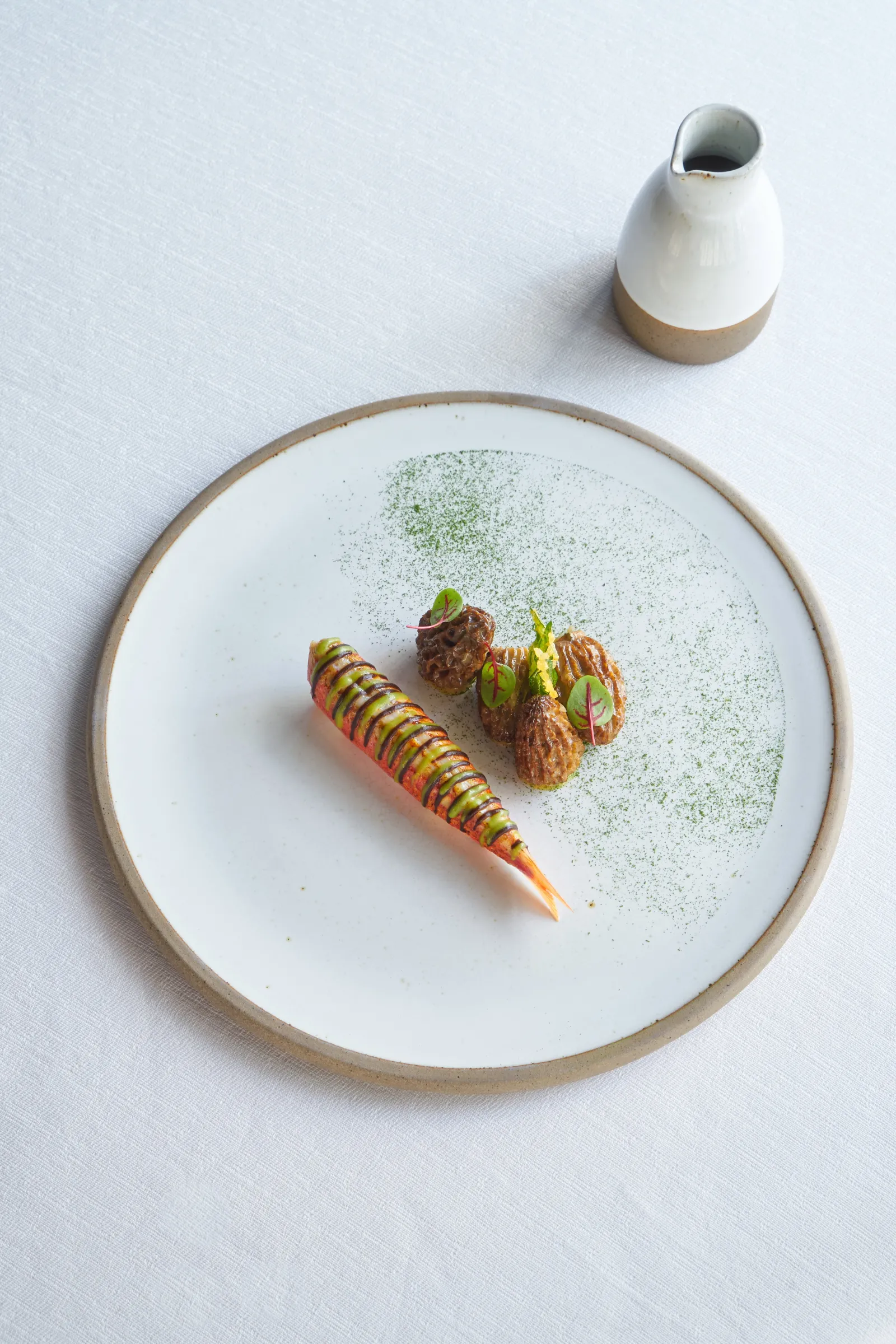 Assiette de présentation gastronomique avec carotte multicolore, croquettes et microherbes vertes, accompagnée d'une petite cruche en céramique blanche et beige