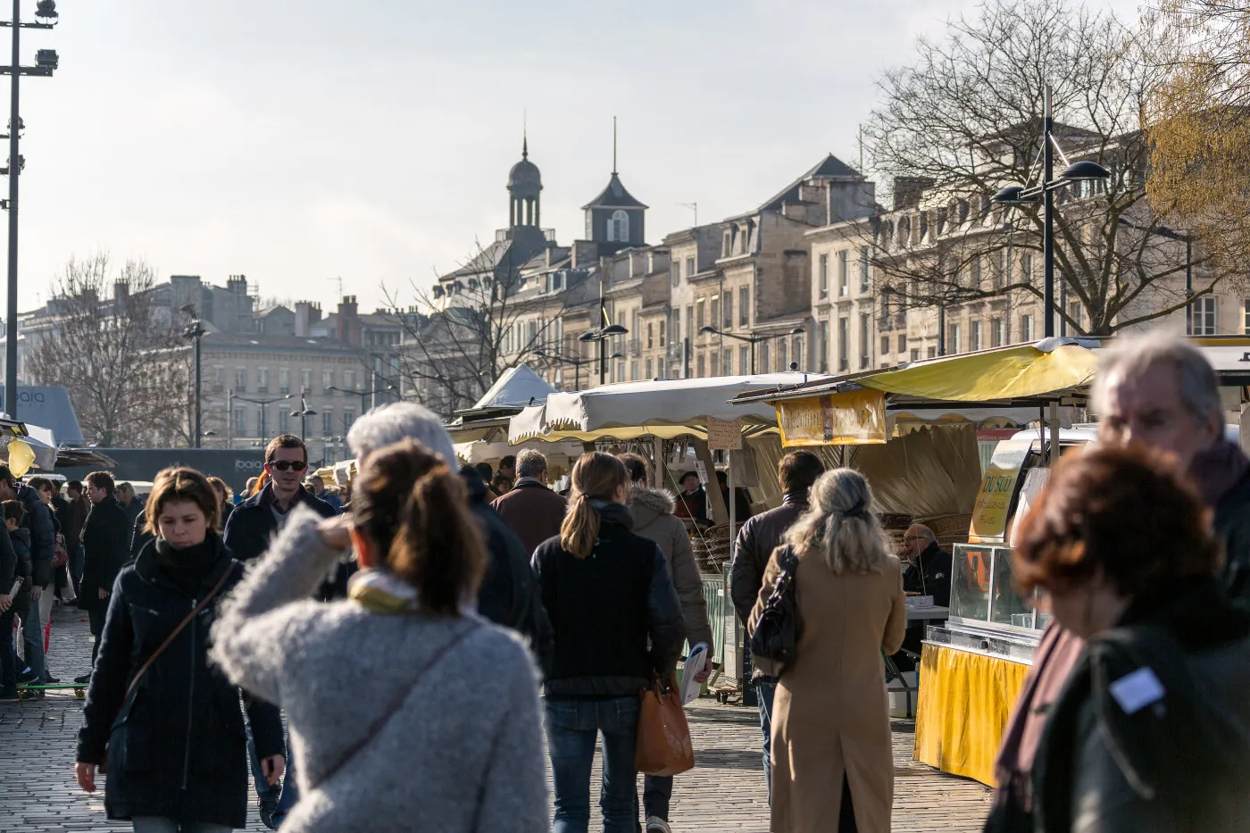 Marché en plein air au quai des Chartrons à Bordeaux avec des étals jaunes, des visiteurs flânant entre les stands, et l'architecture haussmannienne de la ville en arrière-plan sous un ciel dégagé.