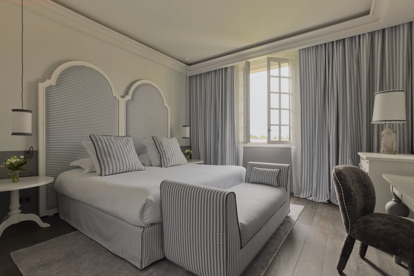 Chambre élégante du Como Cordeillan-Bages à Bordeaux avec lit à rayures grises et blanches, tête de lit arquée, rideaux gris clair, fenêtre donnant sur la vue, bureau avec lampe et chaise noire, décoration minimaliste et raffinée.
