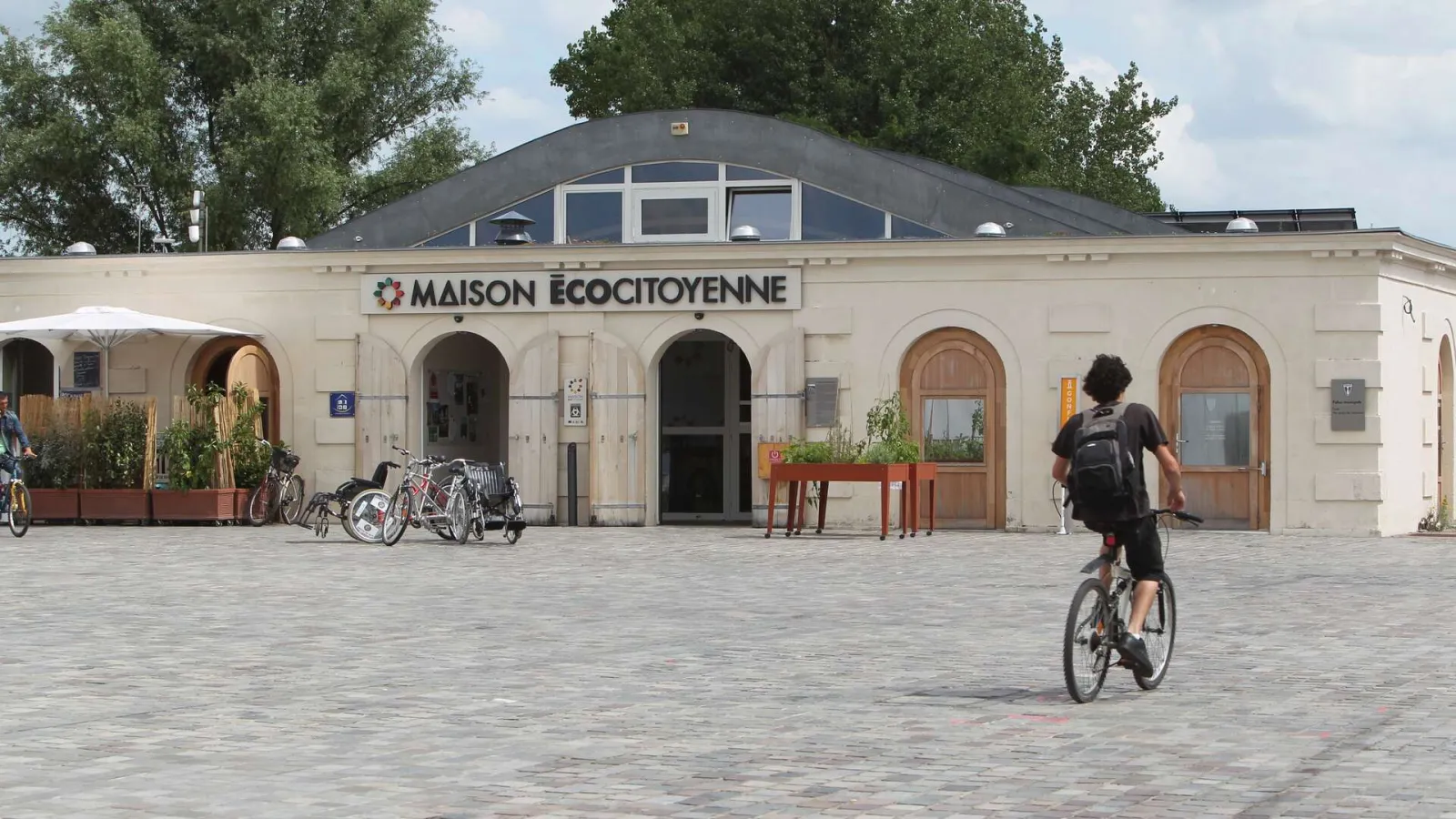 Façade de la Maison écocitoyenne de Bordeaux, bâtiment blanc avec arches, plusieurs vélos garés devant et une personne à vélo sur la place
