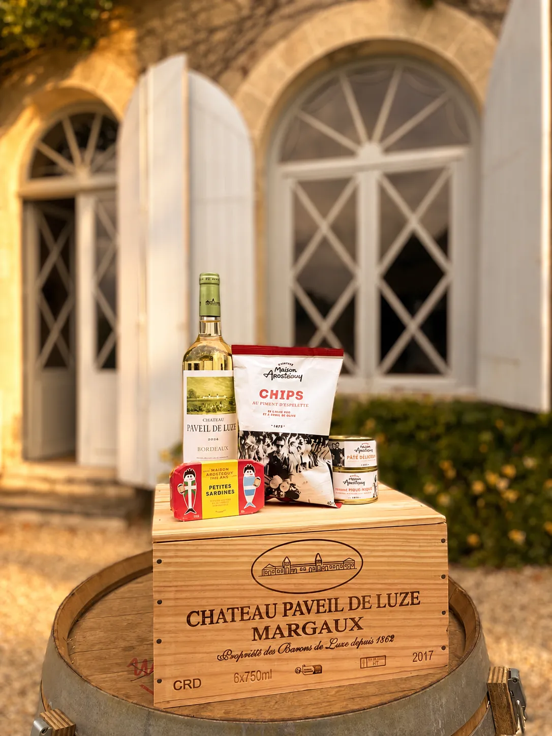 Panier gastronomique du Château Paveil de Luze à Margaux contenant une bouteille de vin blanc, des chips, des crackers et des produits régionaux, présenté sur un tonneau en bois devant une villa bordelaise illuminée