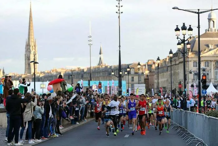 Coureurs participant au semi-marathon de Bordeaux, courant sur une route fermée avec des spectateurs le long du parcours et l'architecture historique de Bordeaux en arrière-plan.