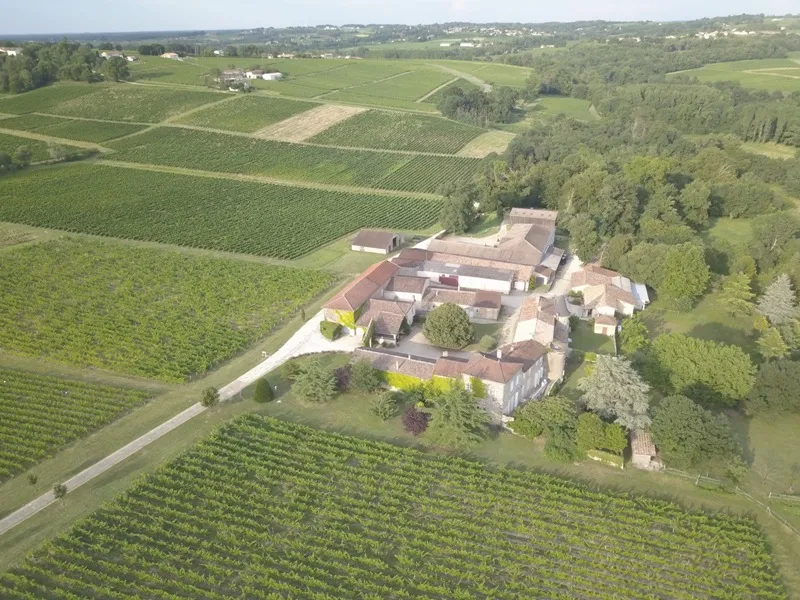 Vue aérienne du Château Monconseil-Gazin à Bordeaux, entouré de vignobles verdoyants et de jardins paysagers, avec ses bâtiments en tuiles roses et ses installations viticoles.