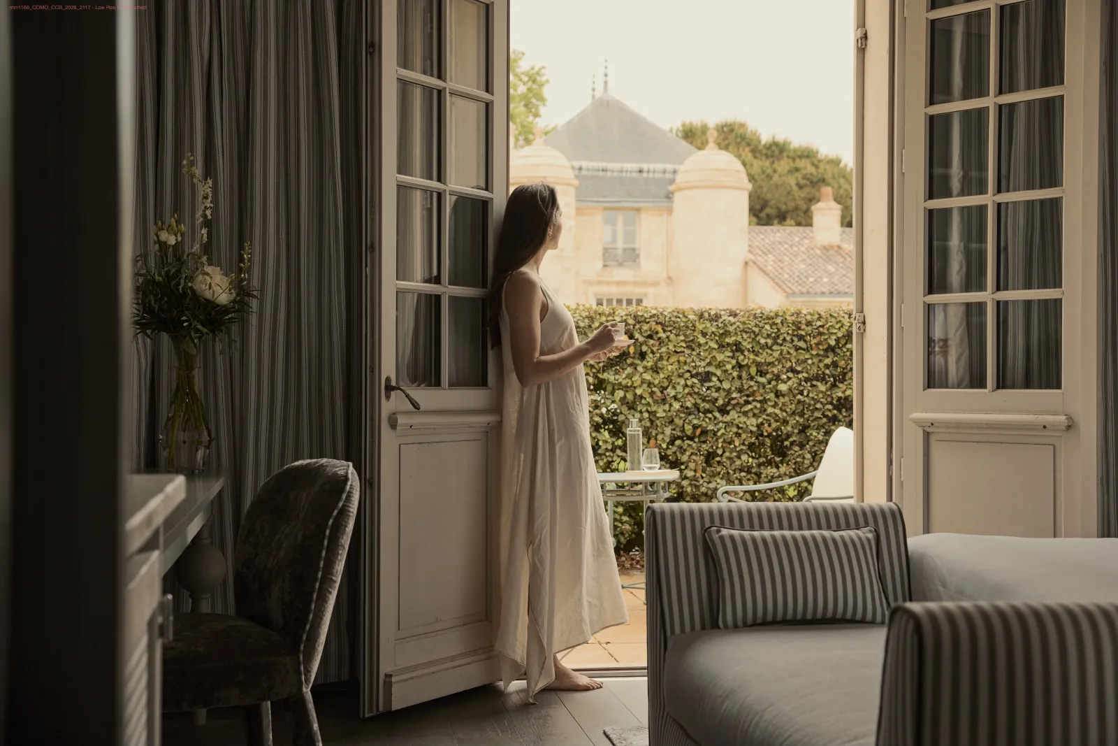 Femme en robe blanche se tenant debout sur une terrasse avec portes ouvertes, regardant vers un château et des vignes à Bordeaux, Como Cordeillan-Bages