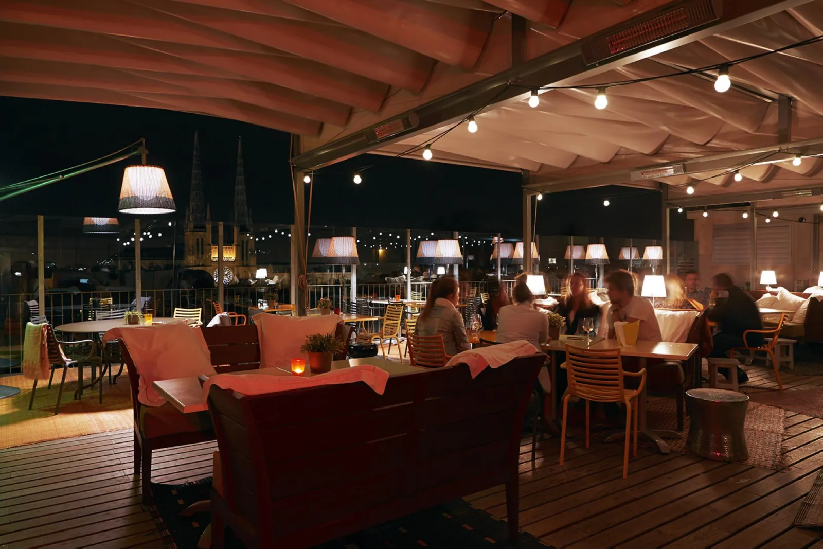 Terrasse extérieure du Mama Shelter Bar à Bordeaux la nuit, avec des clients assis autour de tables, des lampes chauffantes, une structure couverte en bois et des luminaires suspendus.