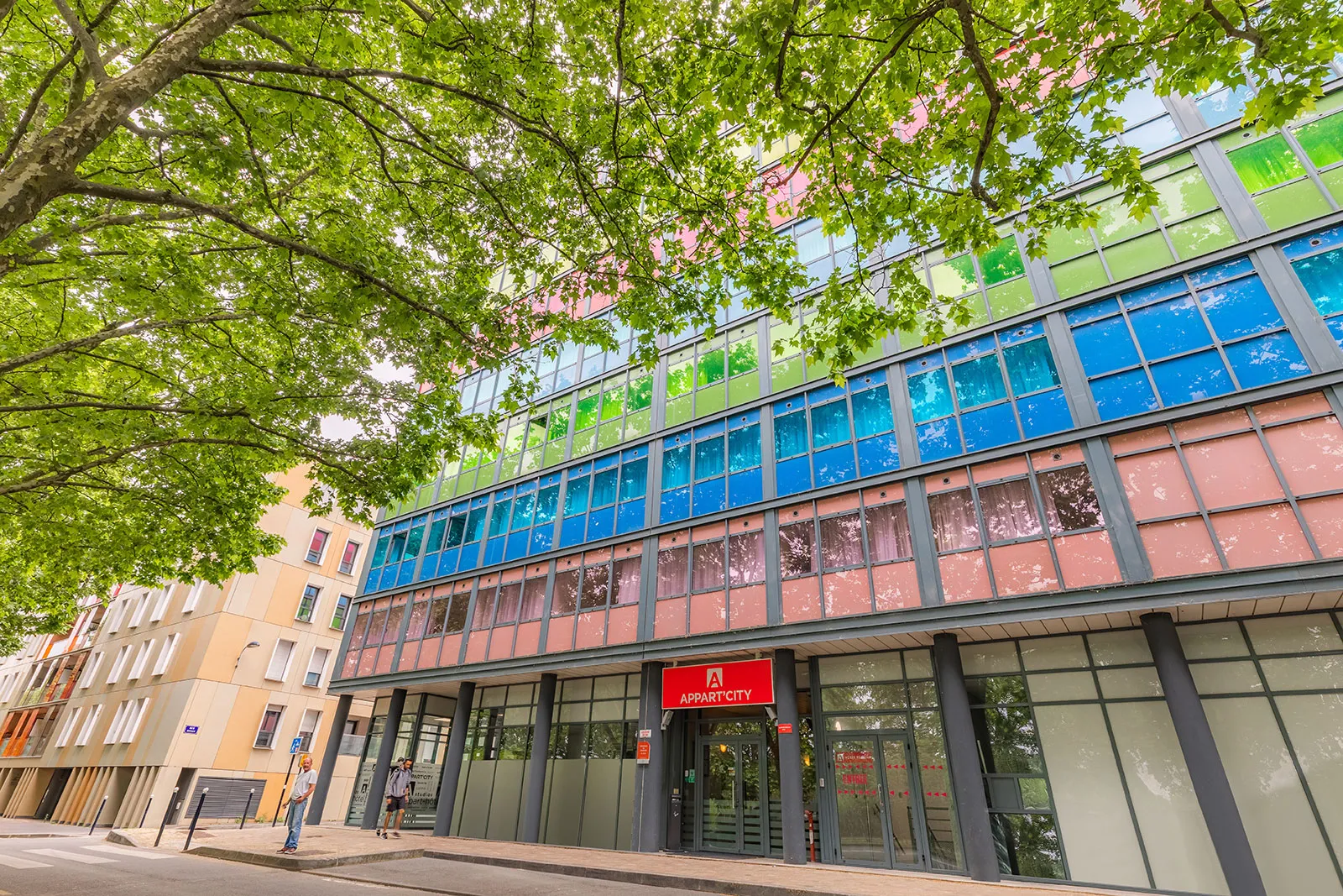 Façade moderne du bâtiment Appart'City Classic à Bordeaux avec des panneaux de verre colorés bleu, vert et rose, ombragée par un grand arbre feuillu, entourée de bâtiments de style haussmannien.