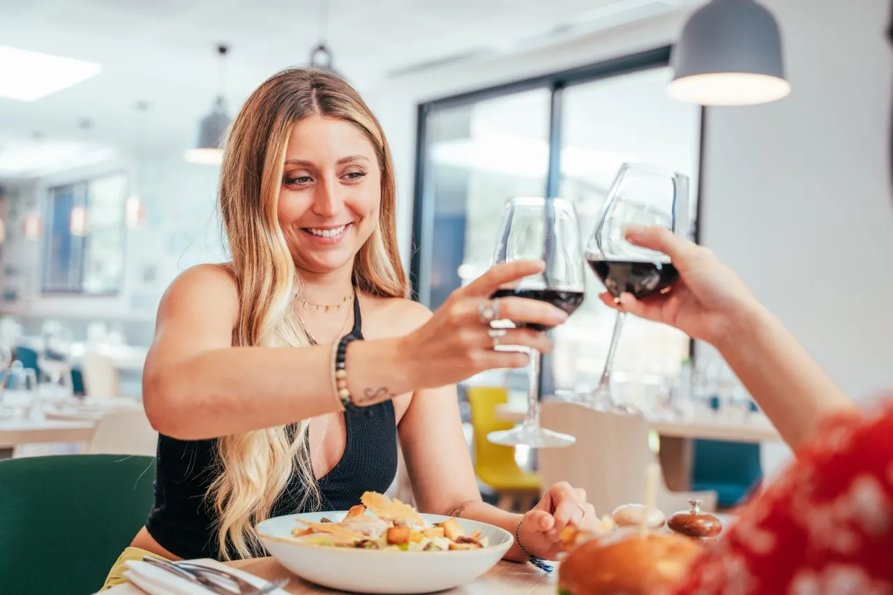 Une femme souriante trinque avec un verre de vin rouge lors d'un repas dans un restaurant moderne de l'Holiday Inn Bordeaux Mérignac, avec une assiette de salade devant elle.