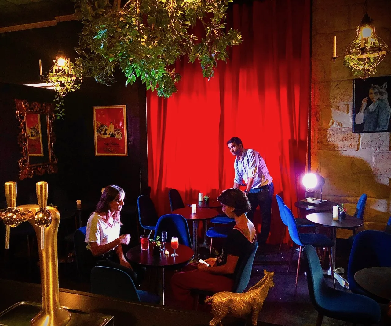 Intérieur du Théâtre des Chartrons à Bordeaux avec deux personnes assises à une table devant des verres, un serveur debout en arrière-plan, rideau rouge dramatique, éclairage chaleureux et décoration vintage.