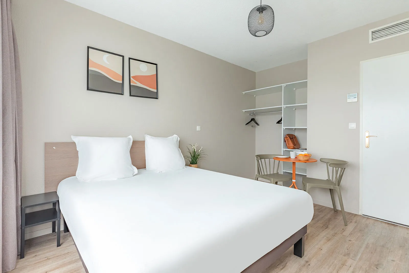 Chambre moderne d'un appartement à Bordeaux avec un lit double aux draps blancs, un bureau avec chaise, un placard ouvert, et une décoration minimaliste aux touches orangées.