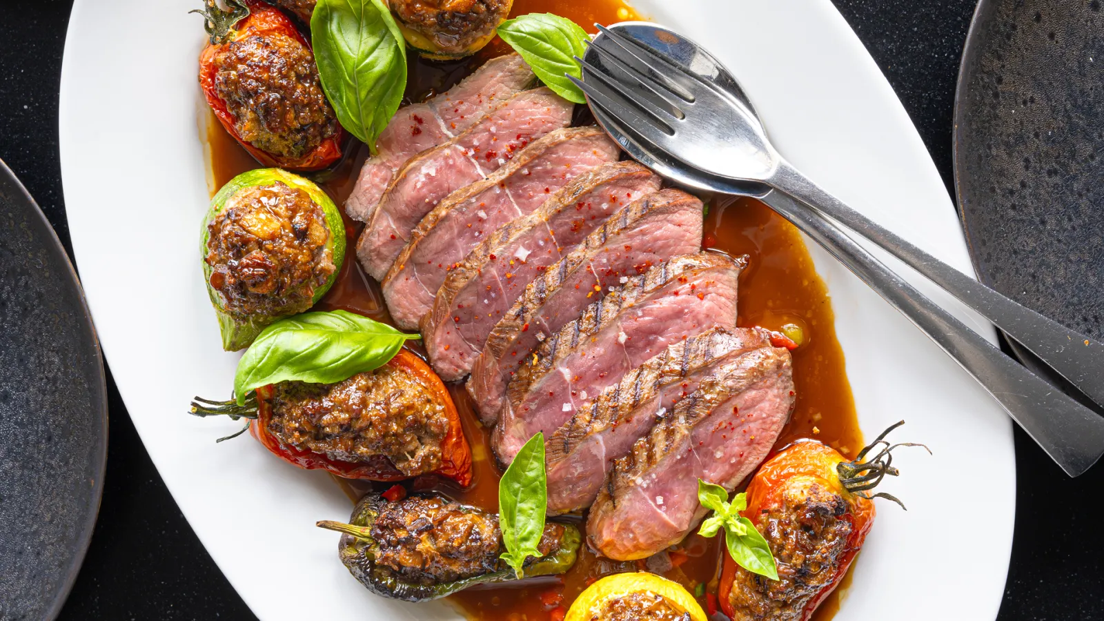 Assiette de viande rouge saignante tranchée accompagnée de tomates et poivrons farcis et grillés, décorée de feuilles de basilic frais, servie avec une sauce au jus.