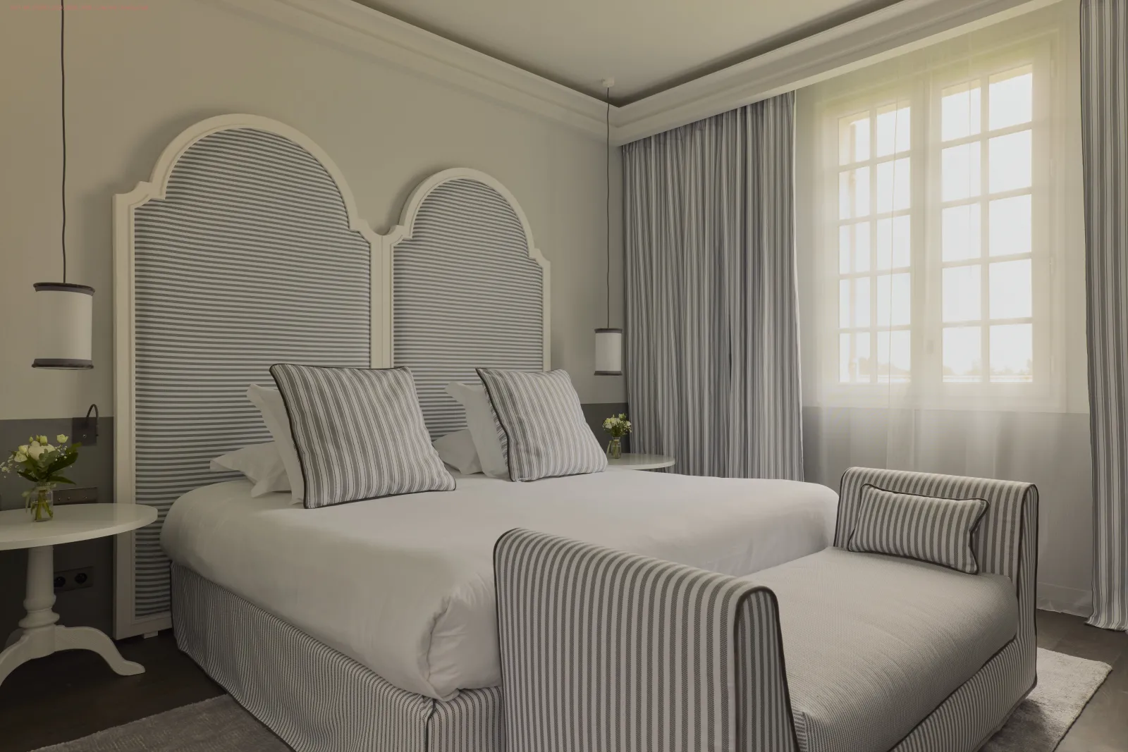 Chambre élégante avec lit king-size à tête de lit arquée striée, oreillers assortis, draps blancs, fenêtres voilées et rideaux gris, décoration minimaliste dans un style classique contemporain.