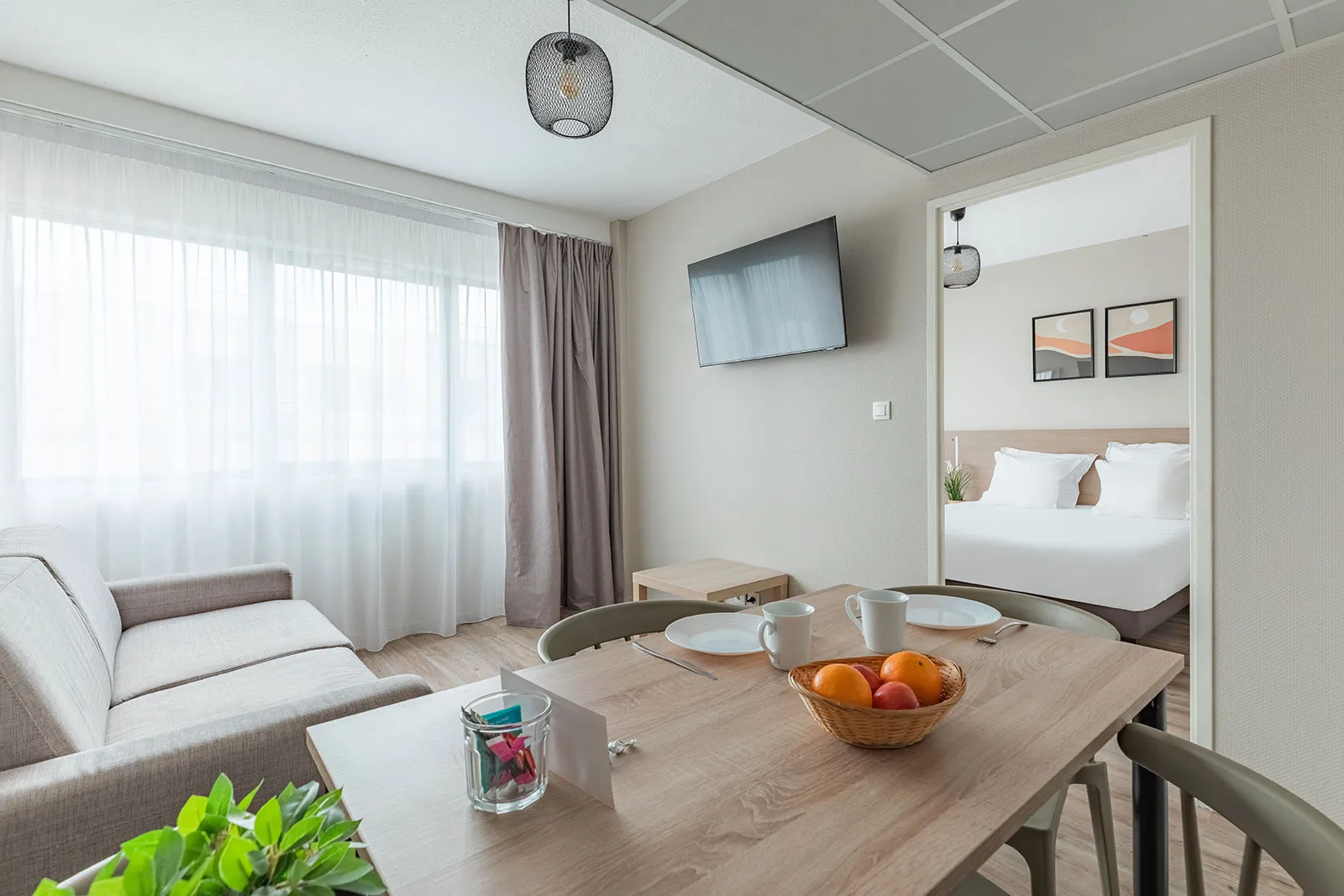 Suite spacieuse d'un appartement-hôtel à Bordeaux avec coin salon, table à manger équipée de fruits frais, accès à la chambre avec lit double, TV murale et grandes baies vitrées avec rideaux.