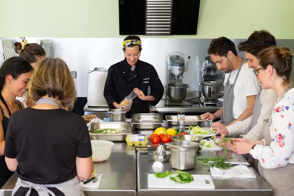 Cours de cuisine à Bordeaux chez Fondant cuisine : un chef en tablier noir explique une recette à un groupe d'adultes autour d'un plan de travail rempli de légumes frais et d'ustensiles de cuisine professionnels.