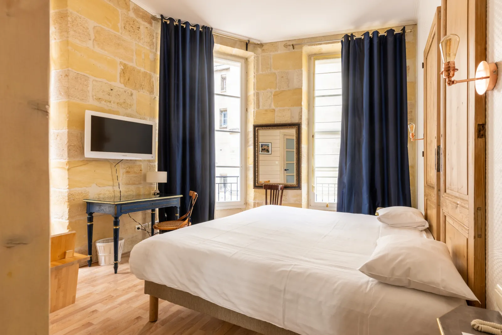 Chambre élégante de l'Hôtel de la Tour Intendance à Bordeaux, avec un lit blanc, murs en pierre dorée, rideaux bleu foncé, bureau avec écran plat et vue sur la ville depuis les fenêtres.