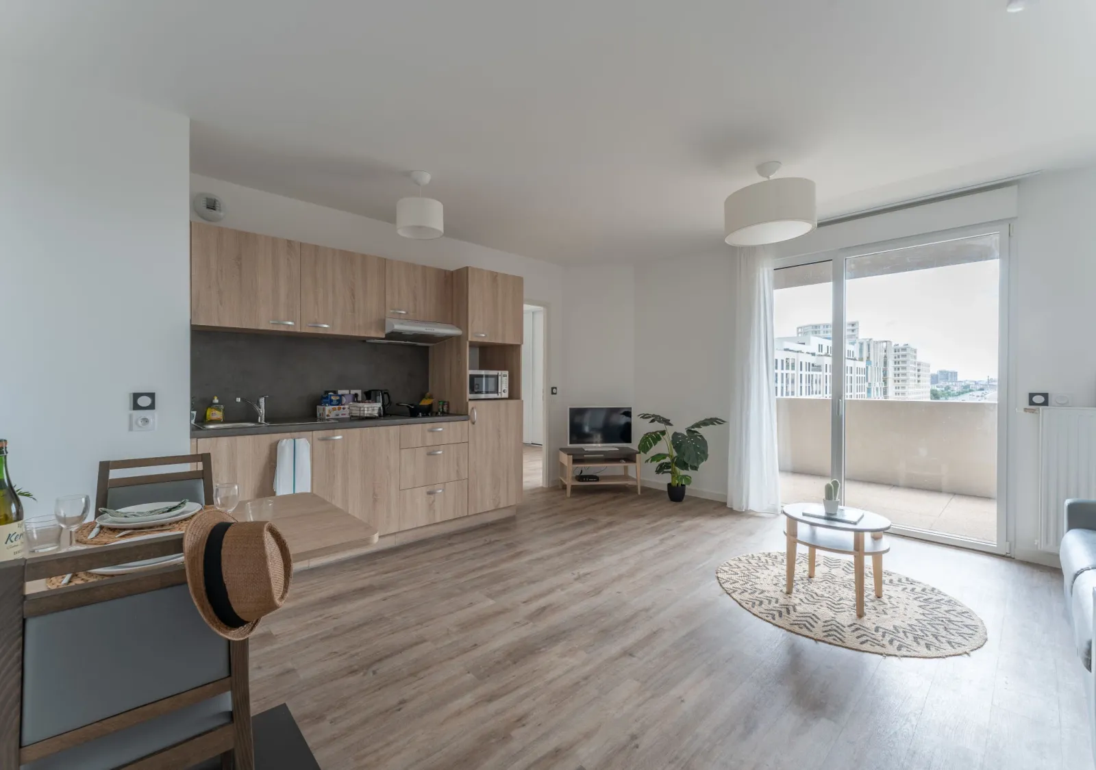 Appartement moderne et lumineux avec cuisine équipée en bois clair, salon avec télévision, table basse et plantes vertes, ouvert sur un balcon avec vue sur la ville de Bordeaux.
