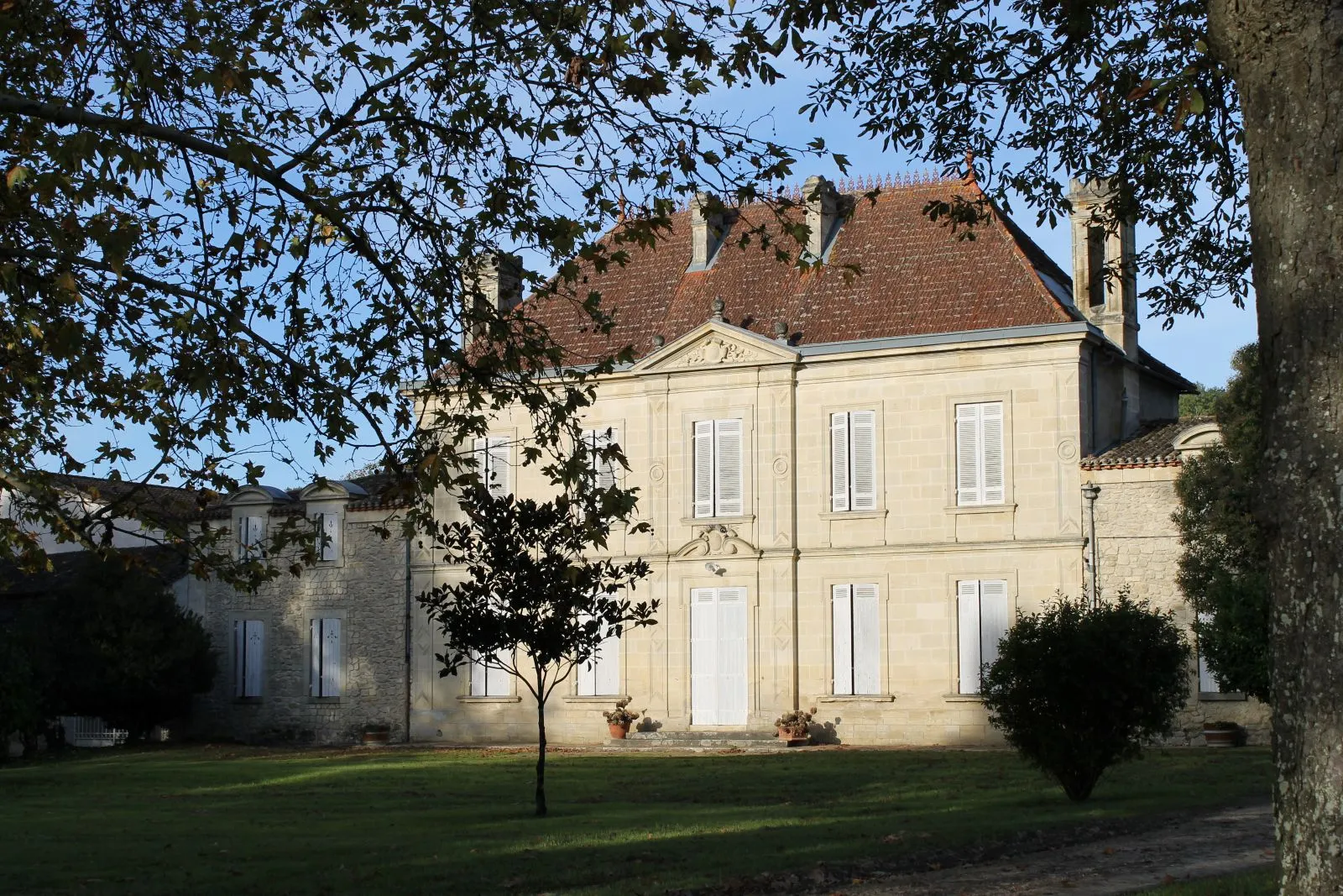 Château Mangot à Bordeaux, une élégante demeure en pierre de taille de couleur crème avec toit en tuiles rouges, entourée d'une pelouse et d'arbres, façade symétrique avec fenêtres blanches et porche central.