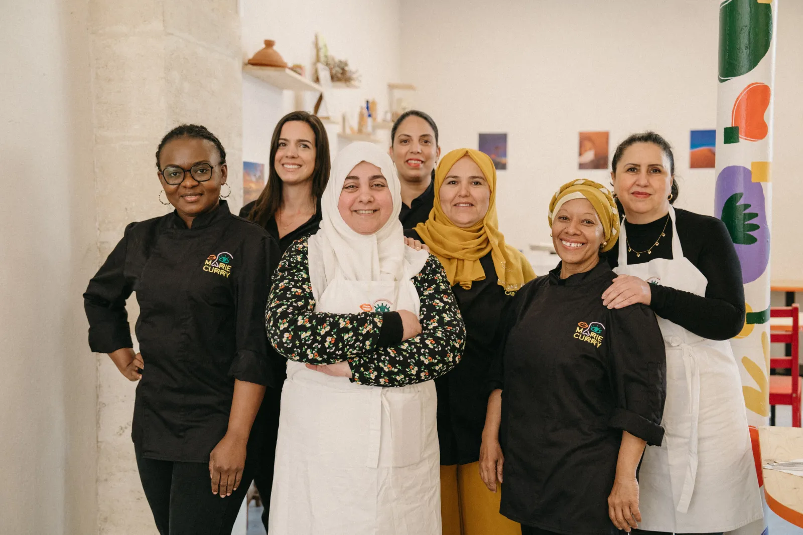 Équipe souriante du restaurant Marie Curry à Bordeaux, composée de six femmes en uniforme noir et tenues traditionnelles colorées, posant ensemble dans la cuisine du restaurant.