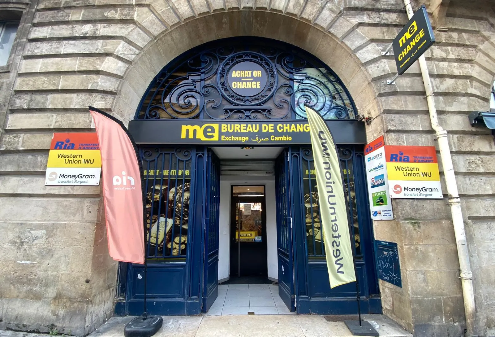 Devanture du bureau de change Monex Exchange situé à Bordeaux, rue Victor Hugo, avec portes bleues ornées de motifs dorés, enseigne jaune et affiches publicitaires Western Union et MoneyGram.
