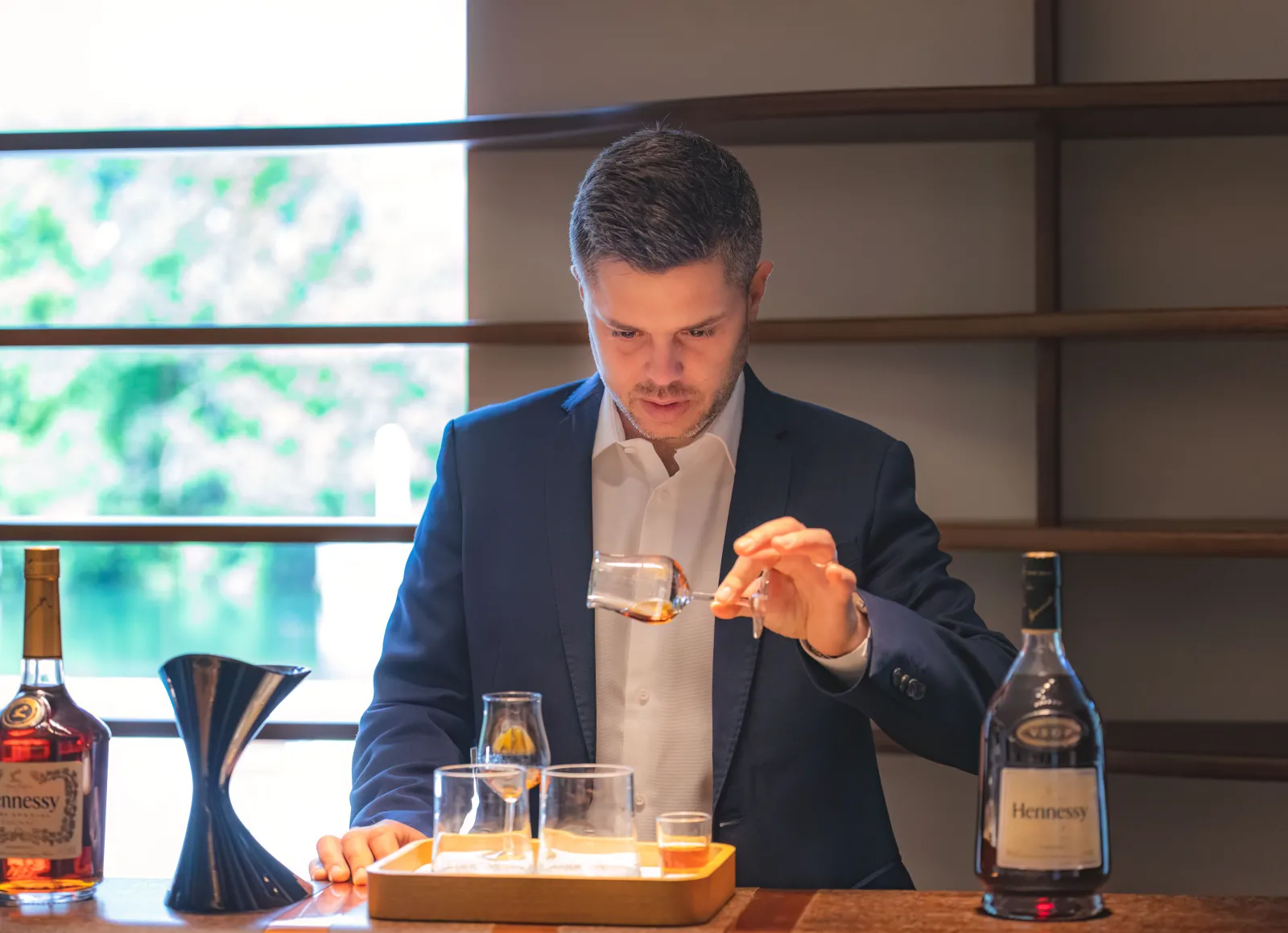 Un homme en costume bleu présentant un verre de cognac Hennessy lors d'une dégustation, avec des bouteilles de Hennessy sur la table et une vue de Bordeaux en arrière-plan.