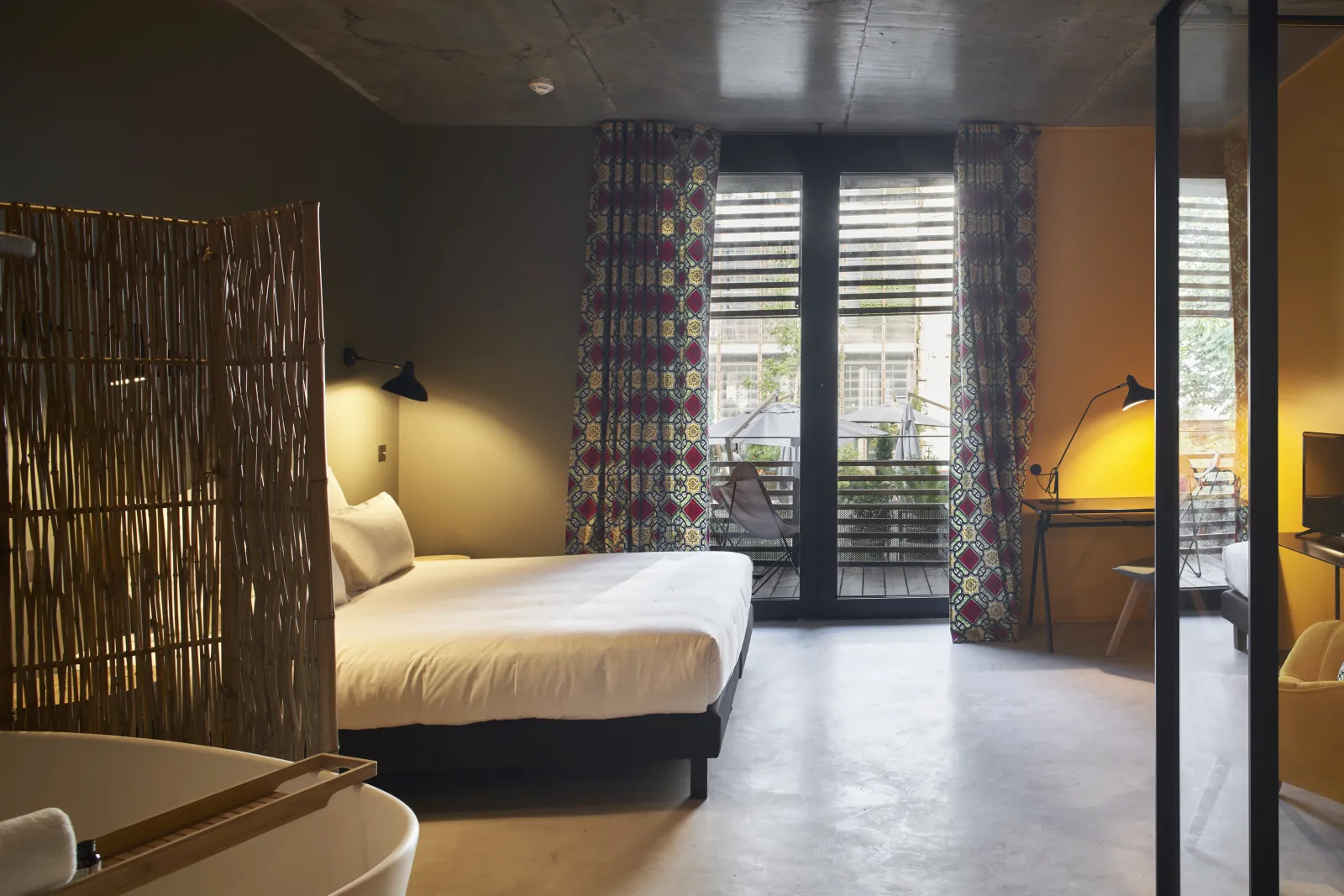 Chambre moderne de l'Hôtel La Zoologie à Bordeaux avec lit blanc, cloison en bois, murs jaunes, portes-fenêtres donnant sur balcon et rideaux à motifs colorés