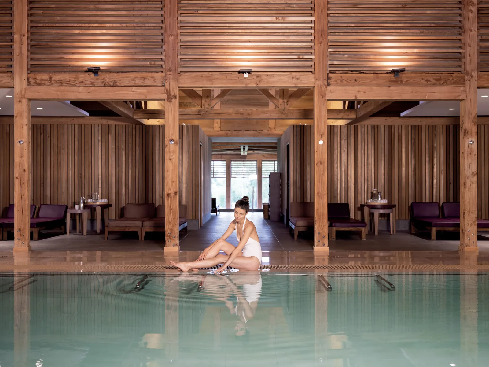 Une femme assise au bord d'une piscine intérieure dans un spa aux architectures en bois massif, avec des espaces de détente aménagés en arrière-plan.
