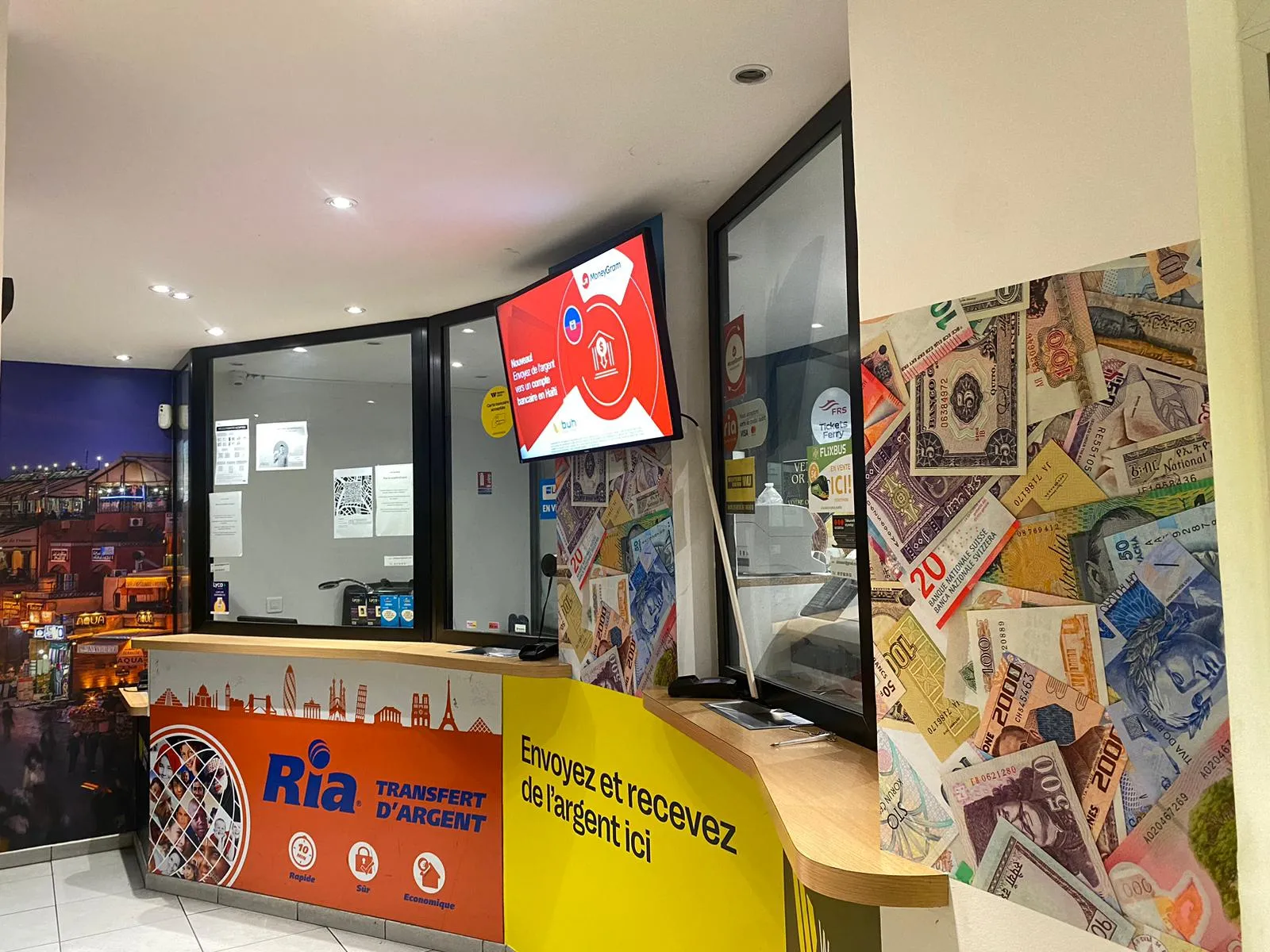 Devanture du bureau de change Ria Transfert d'Argent à Bordeaux Gambetta, avec comptoir de service, affichage numérique et décoration murale représentant des billets de banque et des monuments parisiens.