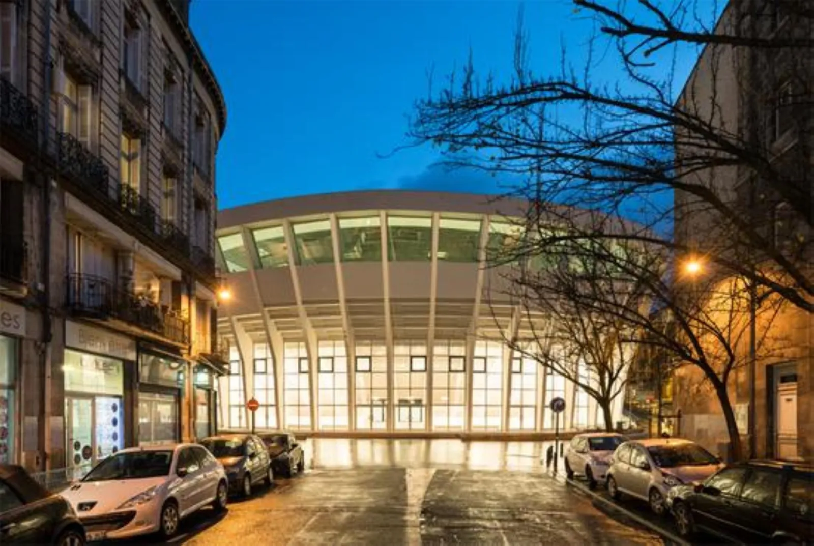 Vue nocturne du Palais des Sports de Bordeaux avec sa façade moderne en verre illuminée, entouré de bâtiments haussmanniens et de voitures garées en rue