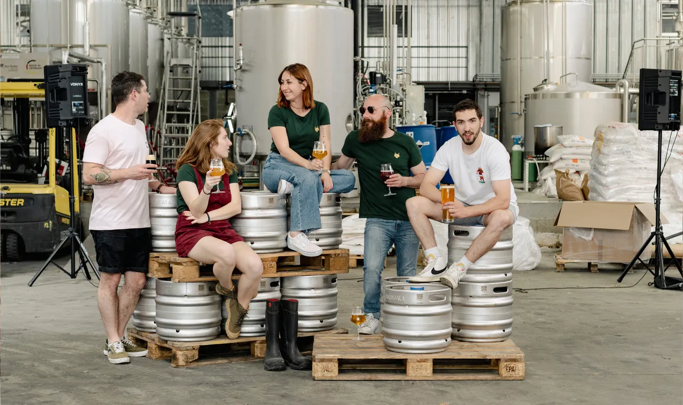 Cinq personnes dégustent de la bière assis sur des fûts en métal dans une brasserie, entourés de réservoirs de fermentation et d'équipements de brassage.