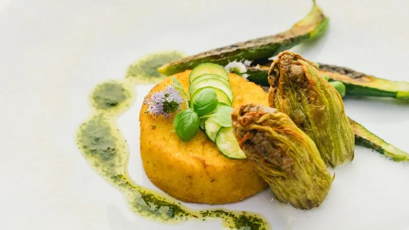 Assiette gastronomique présentant une courge dorée accompagnée de fleurs de courgette farcies, décorée de tranches de concombre, de sauce verte et de microherbes