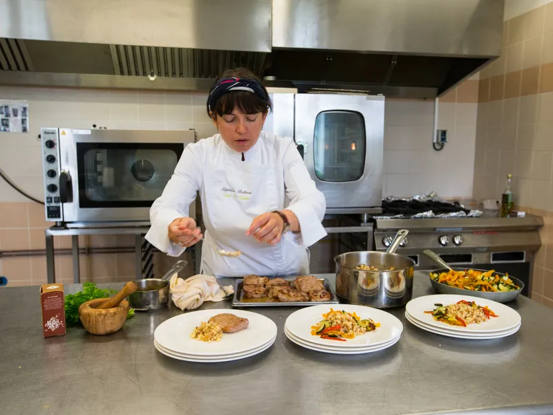 Une chef cuisinière en tenue blanche dresse des assiettes de plats gastronomiques dans la cuisine professionnelle du Château Kirwan à Bordeaux, avec des équipements culinaires et des ingrédients visibles en arrière-plan.