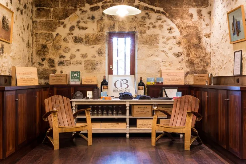 Salle de dégustation du Château Paveil de Luze à Bordeaux, avec murs en pierre ancienne, deux fauteuils Adirondack en bois, comptoir de présentation de vins, et portes en bois massif.