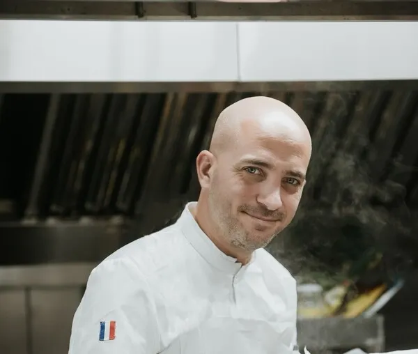 Portrait d'un chef cuisinier chauve souriant, vêtu d'une veste de chef blanche avec un insigne tricolore français, posant dans la cuisine du restaurant Passage Secret à Bordeaux.