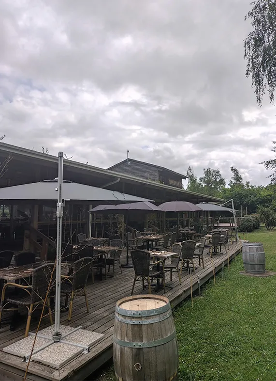 Terrasse couverte du Bistrot du Lac à Bordeaux avec tables et chaises sous des parasols, tonneaux de vin en décoration, et vue sur un jardin verdoyant sous un ciel nuageux