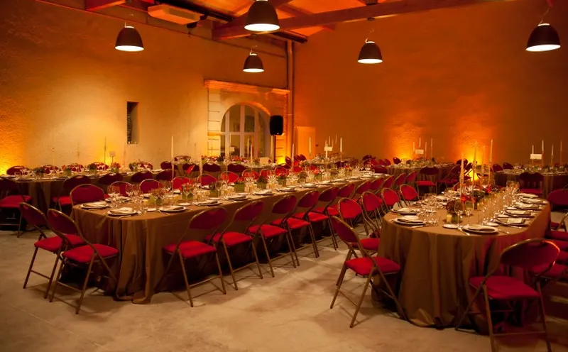 Salle de réception élégante au Château Paveil de Luze à Bordeaux, avec tables rondes recouvertes de tissus dorés, chaises rouges, décoration florale rouge et blanc, éclairage ambiant orange chaud, et plafond aux poutres apparentes.