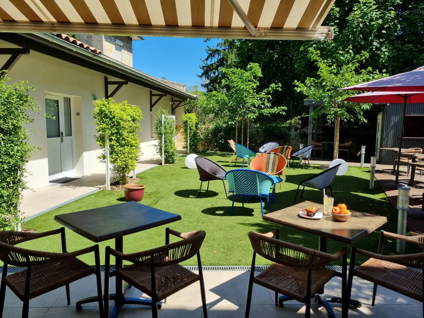 Terrasse extérieure ensoleillée avec pelouse verte, mobilier coloré, tables de pique-nique et parasols rouges, entourée de végétation et d'une maison blanche à Bordeaux