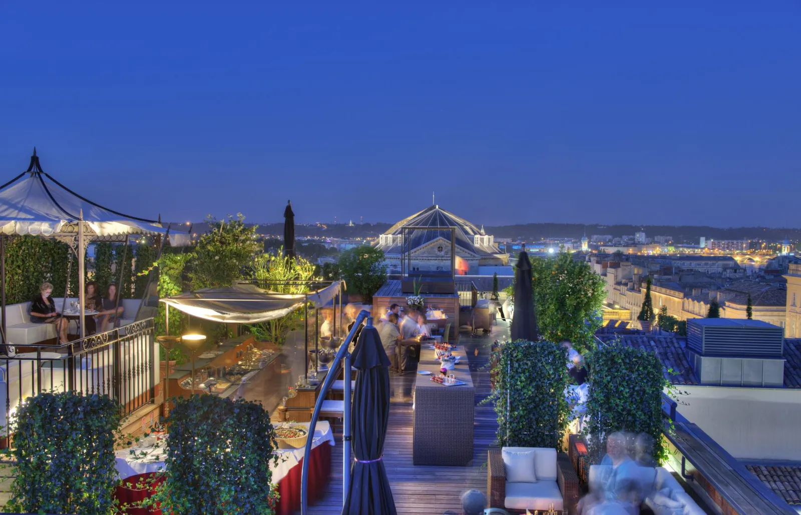 Terrasse panoramique illuminée de l'Intercontinental Grand Hôtel de Bordeaux au crépuscule, avec vue sur la ville et ses monuments historiques, décorée de plantes vertes et de mobilier élégant.