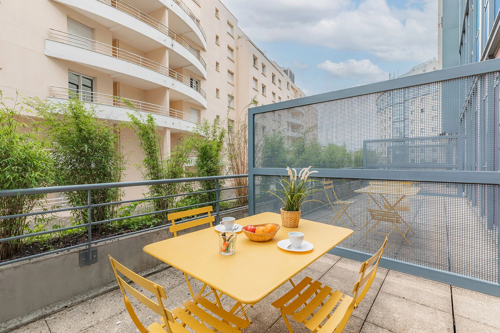 Terrasse ensoleillée d'un appartement à Bordeaux avec table et chaises jaunes, vue sur immeuble résidentiel et jardinière de plantes vertes