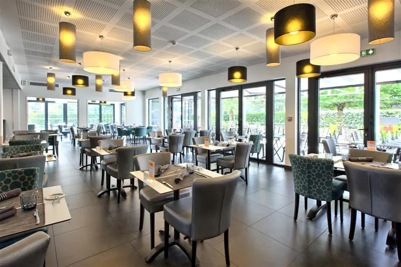Intérieur moderne d'un restaurant avec tables dressées, chaises grises, luminaires suspendus design et grandes baies vitrées donnant sur un jardin verdoyant à Bordeaux.