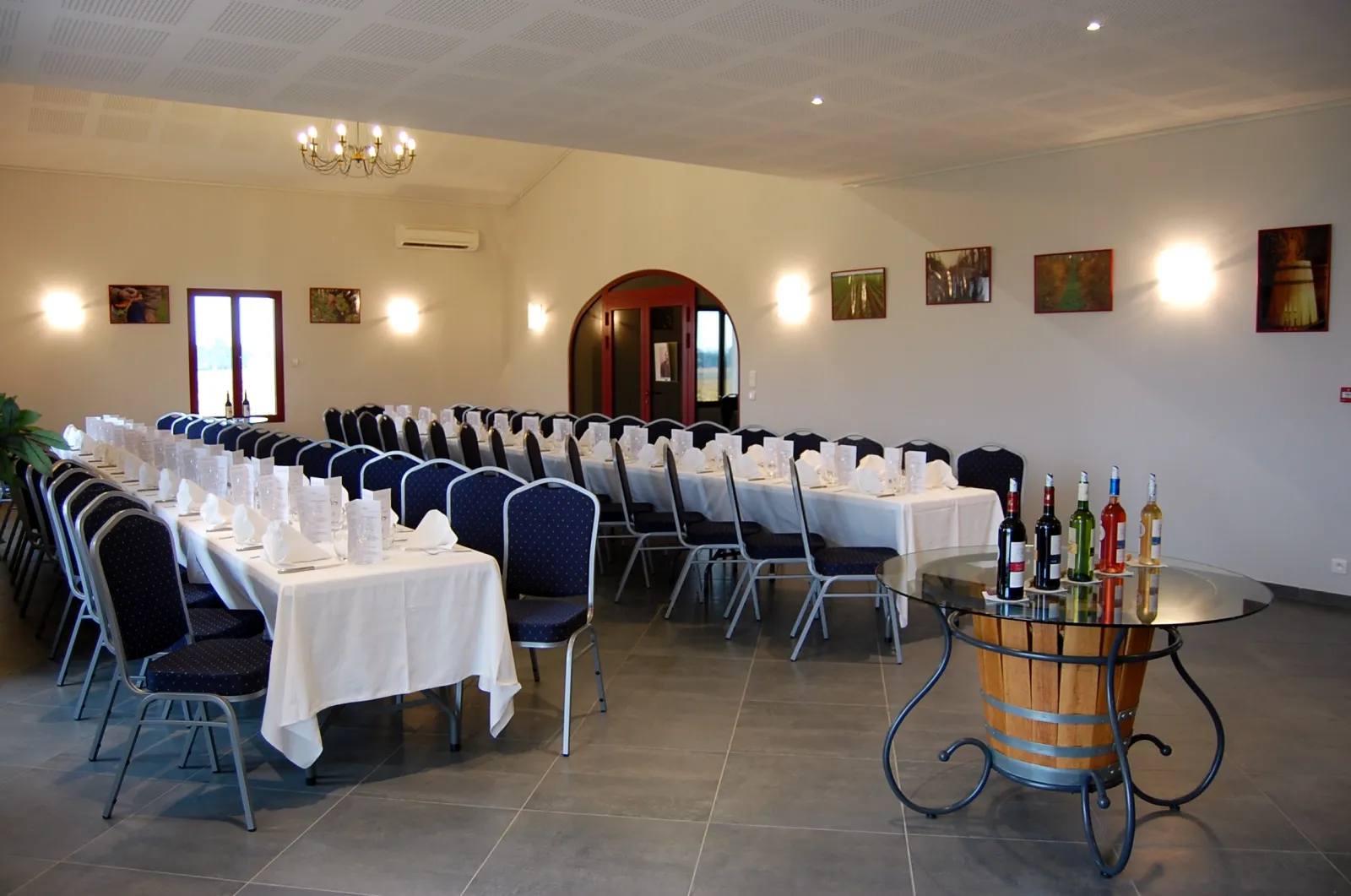 Salle de réception élégante du Château Pichon Bellevue à Bordeaux avec tables dressées en blanc, chaises bleues et noires, tonneau de vin décoratif, et bouteilles de vin disposées sur un présentoir.