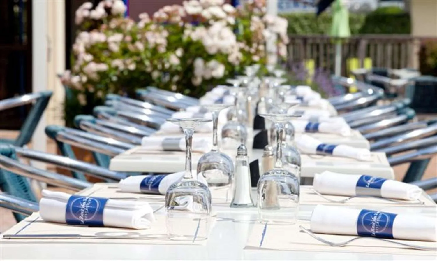 Table de conférence élégante dressée en extérieur avec nappes blanches, verres transparents, serviettes bleues et blanches, fleurs blanches en arrière-plan et chaises bleues