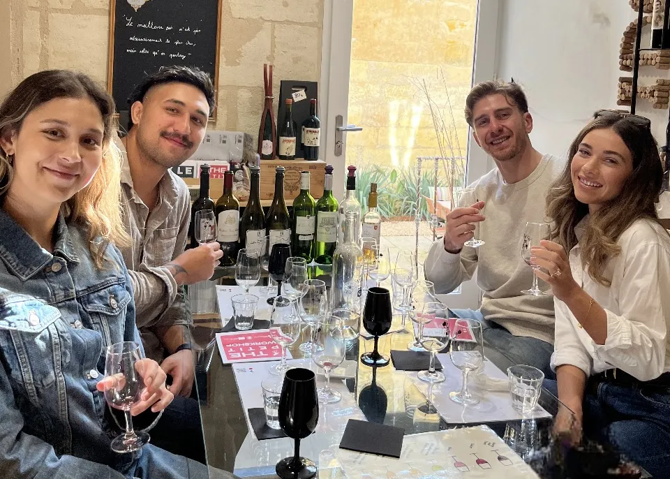 Quatre jeunes adultes souriants lors d'une dégustation de vins à Bordeaux, assis autour d'une table avec plusieurs bouteilles et verres de vin.
