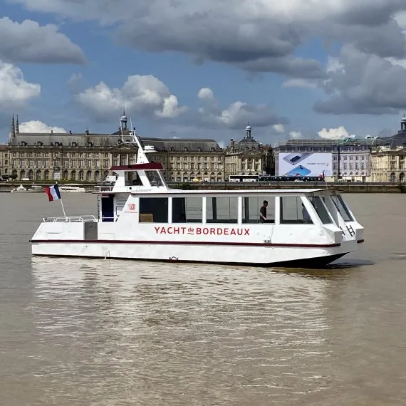 Le Yacht de Bordeaux, bateau de croisière blanc et noir, navigue sur la Garonne avec l'architecture classique de Bordeaux en arrière-plan sous un ciel nuageux.
