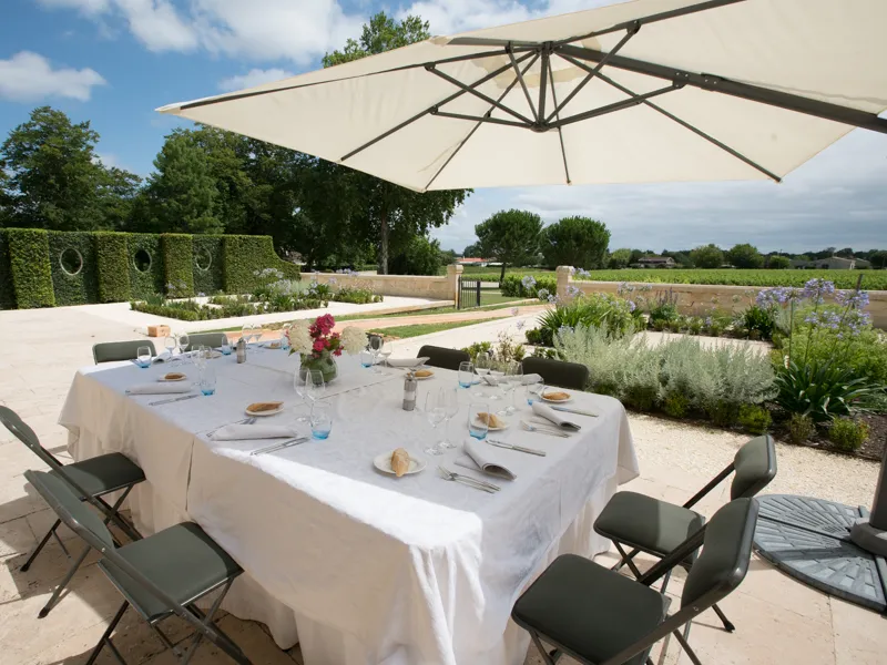 Table dressée en plein air sous un parasol beige au Château Kirwan à Bordeaux, avec couverts, verres et fleurs fraîches, entourée de chaises noires, dans un jardin paysager avec vignobles en arrière-plan.