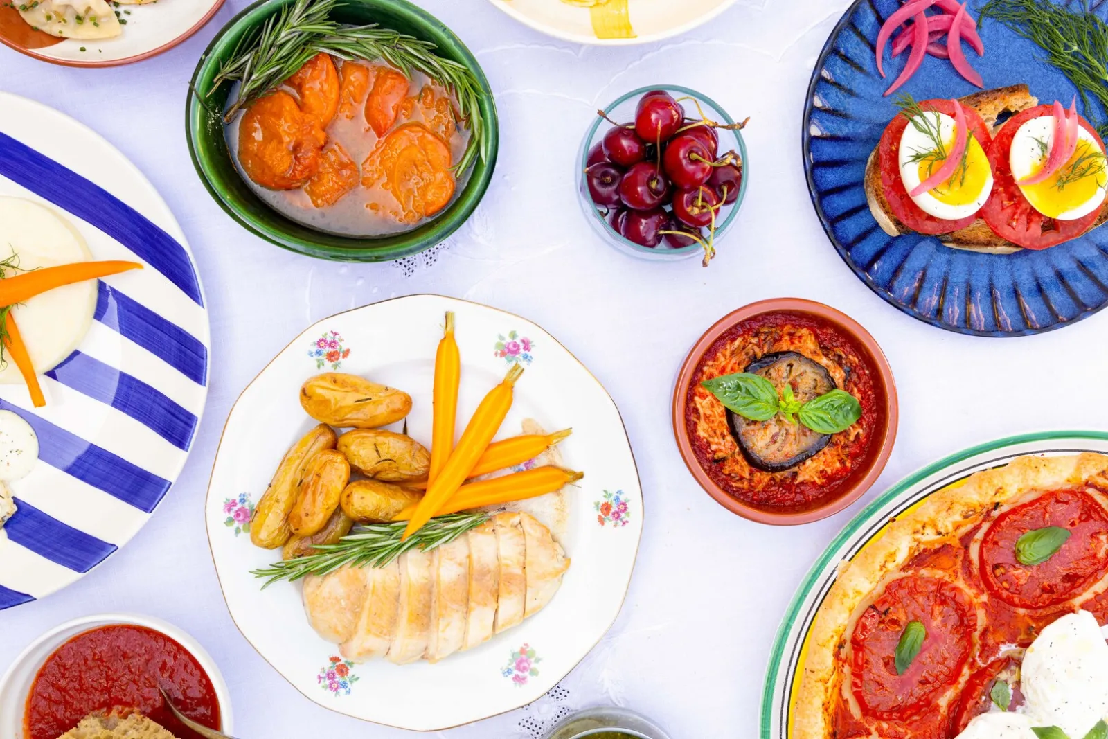 Assortiment de plats cuisinés incluant du poisson avec légumes, des tomates farcies, du pain grillé aux œufs, des fruits frais et des plats traditionnels présentés sur vaisselle colorée
