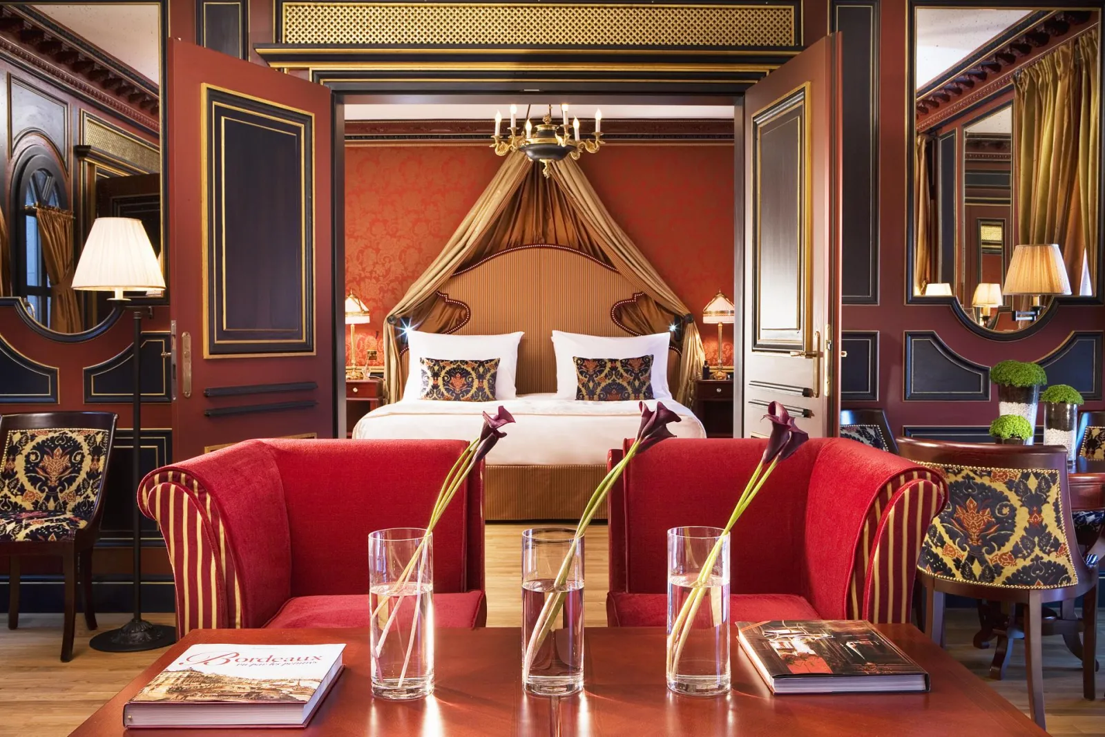 Suite de luxe de l'Intercontinental Grand Hôtel de Bordeaux avec lit à baldaquin doré, murs rouge bordeaux, mobilier classique rouge et or, miroirs ornementaux et éclairage élégant.