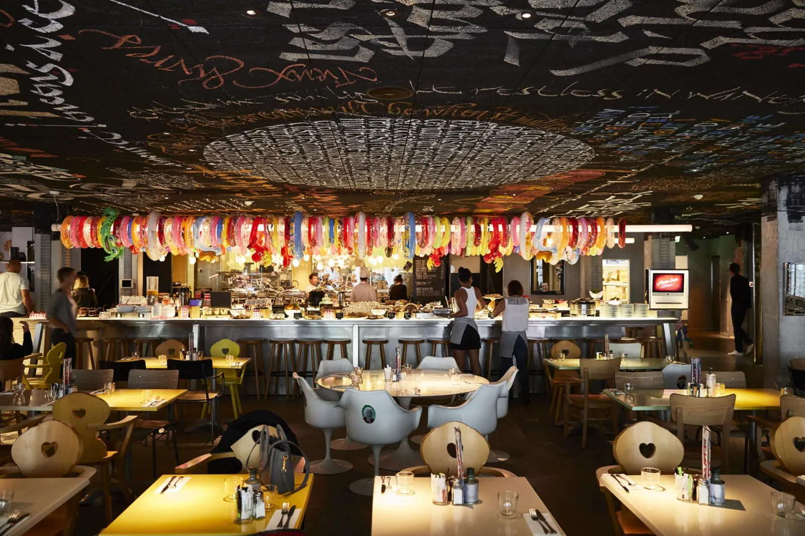 Restaurant Mama Shelter à Bordeaux avec décoration colorée de lanternes suspendues au plafond, tables élégantes avec chaises blanches et jaunes, bar en arrière-plan et ambiance chaleureuse.