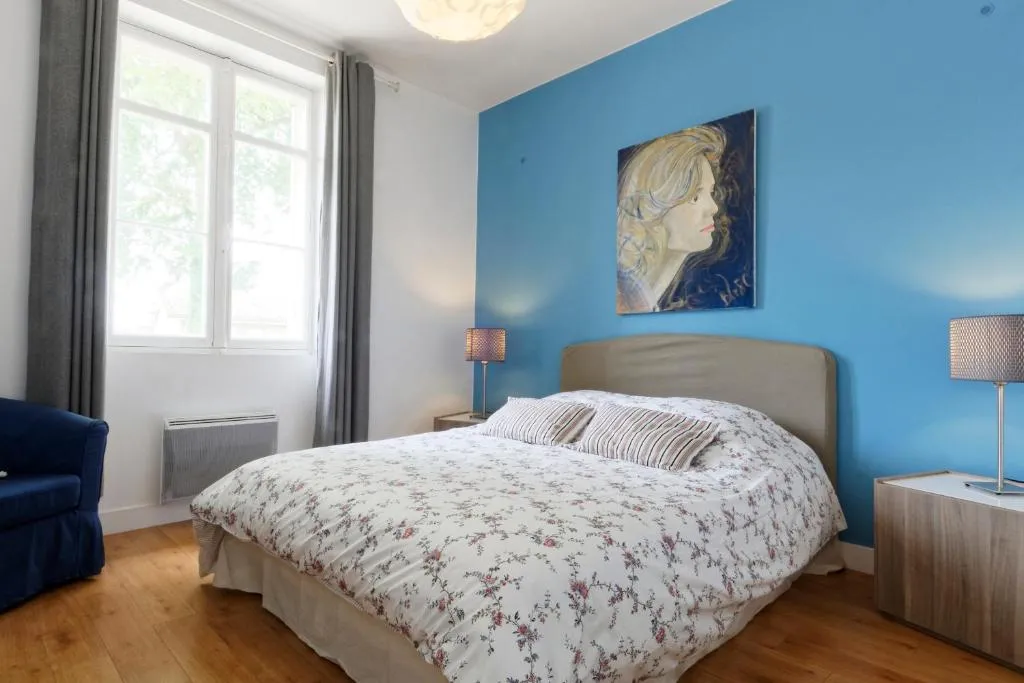 Chambre moderne avec murs bleus, lit queen-size avec linge fleuri, fenêtre donnant sur la verdure, parquet en bois, tableau portrait et mobilier contemporain à Bordeaux